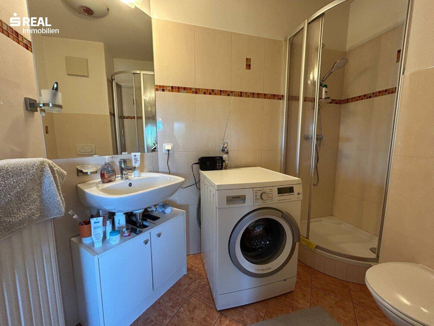 Badezimmer mit Dusche, Waschbecken und Waschmaschine, hell gefliest und praktisch eingerichtet.