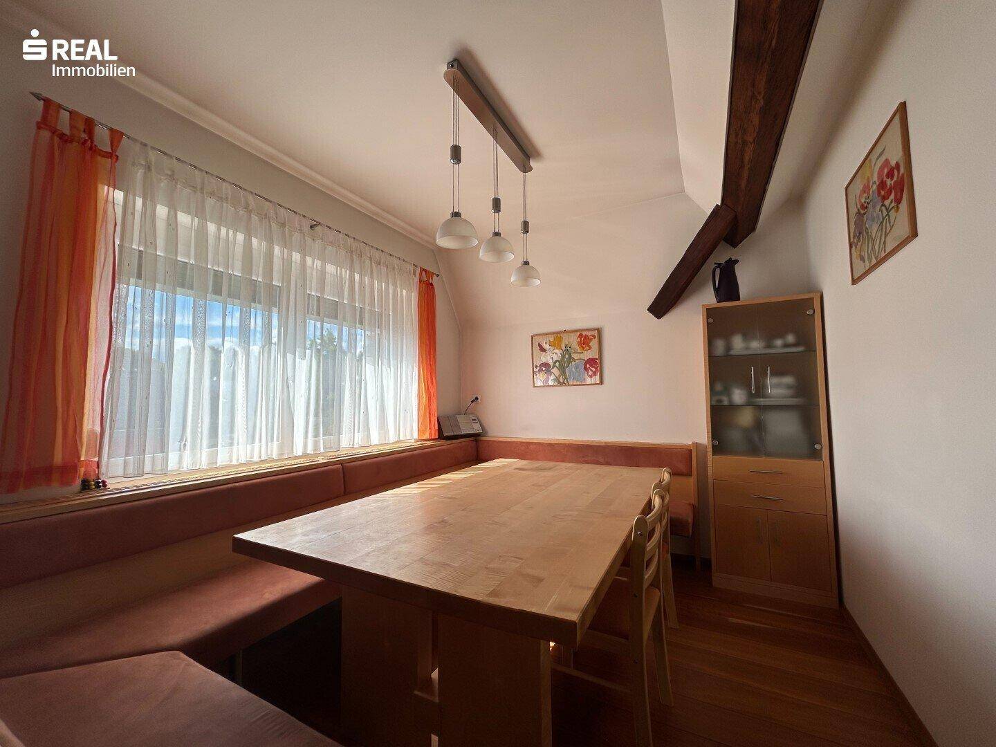 Gemütlicher Essbereich mit Holztisch, Eckbank und großen Fenstern, unter Holzbalken.