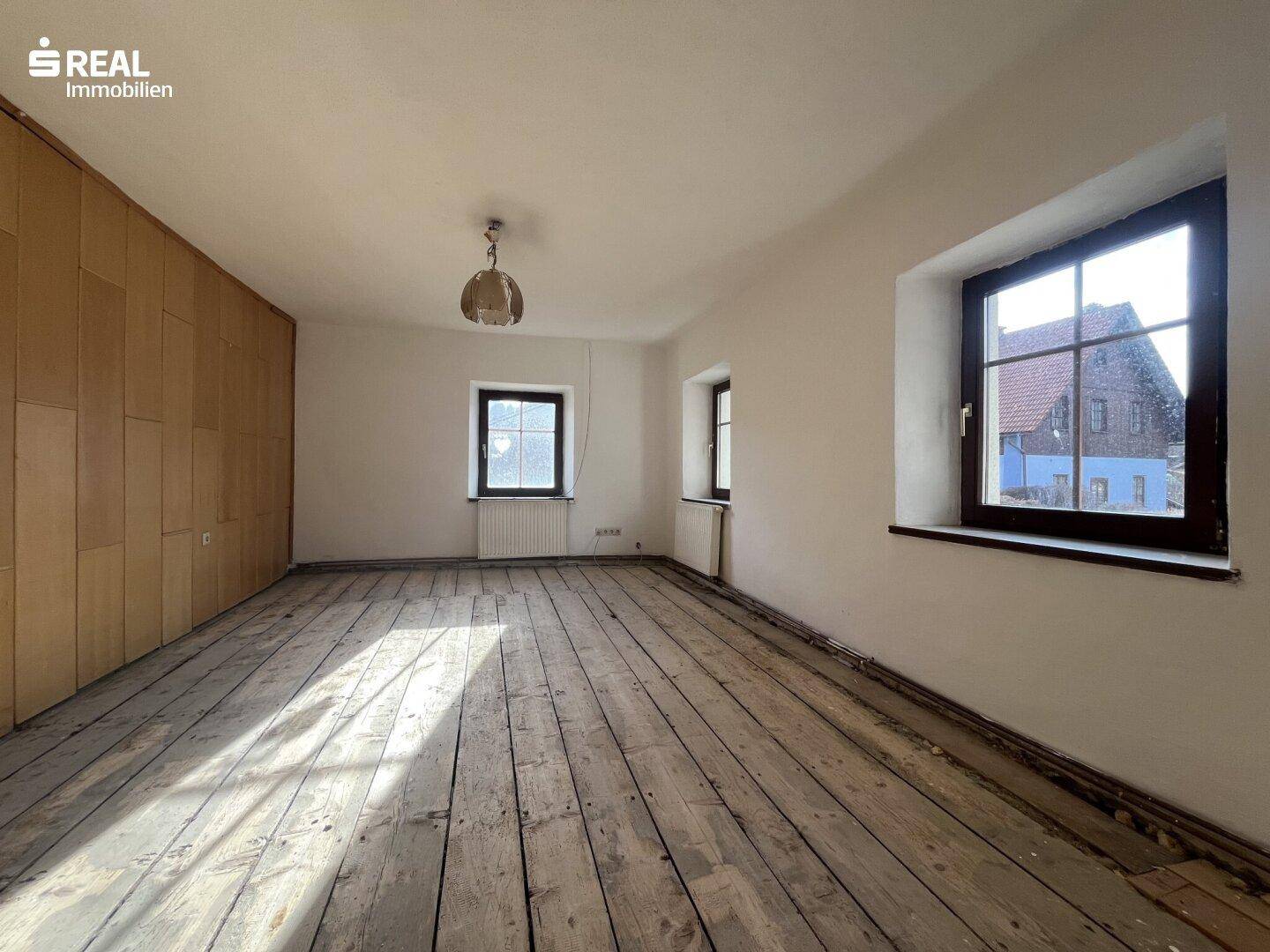 Leerer Raum mit alten Holzplankenböden, weißen Wänden und traditionellen Fenstern.
