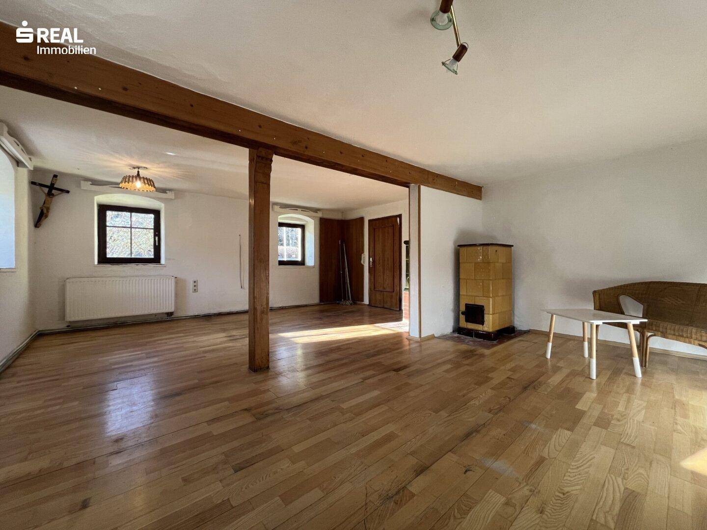 Geräumiger Wohnbereich mit Holzboden, sichtbaren Holzbalken und einem traditionellen Kachelofen.