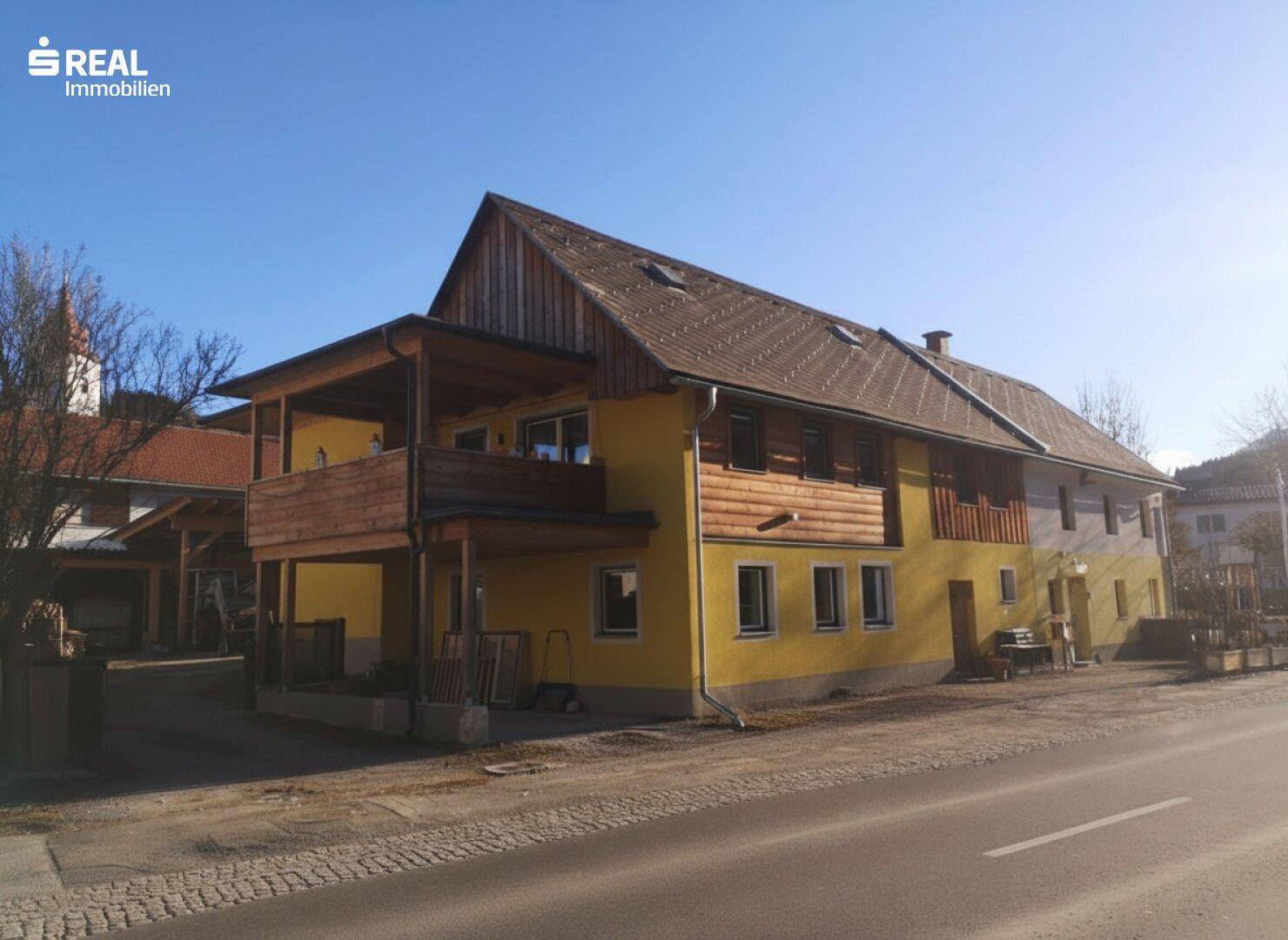 Gebäude mit gelber Fassade und Holzbalkon an einem sonnigen Wintertag in ländlicher Umgebung.