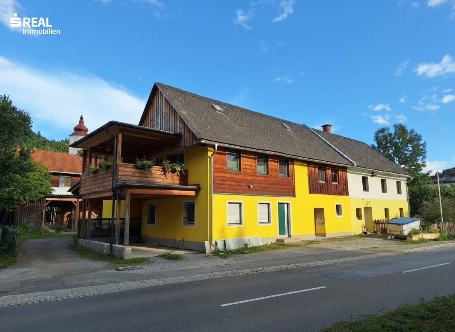 Traditionelles Gebäude mit gelber Fassade, Holzbalkon und Satteldach unter blauem Himmel.