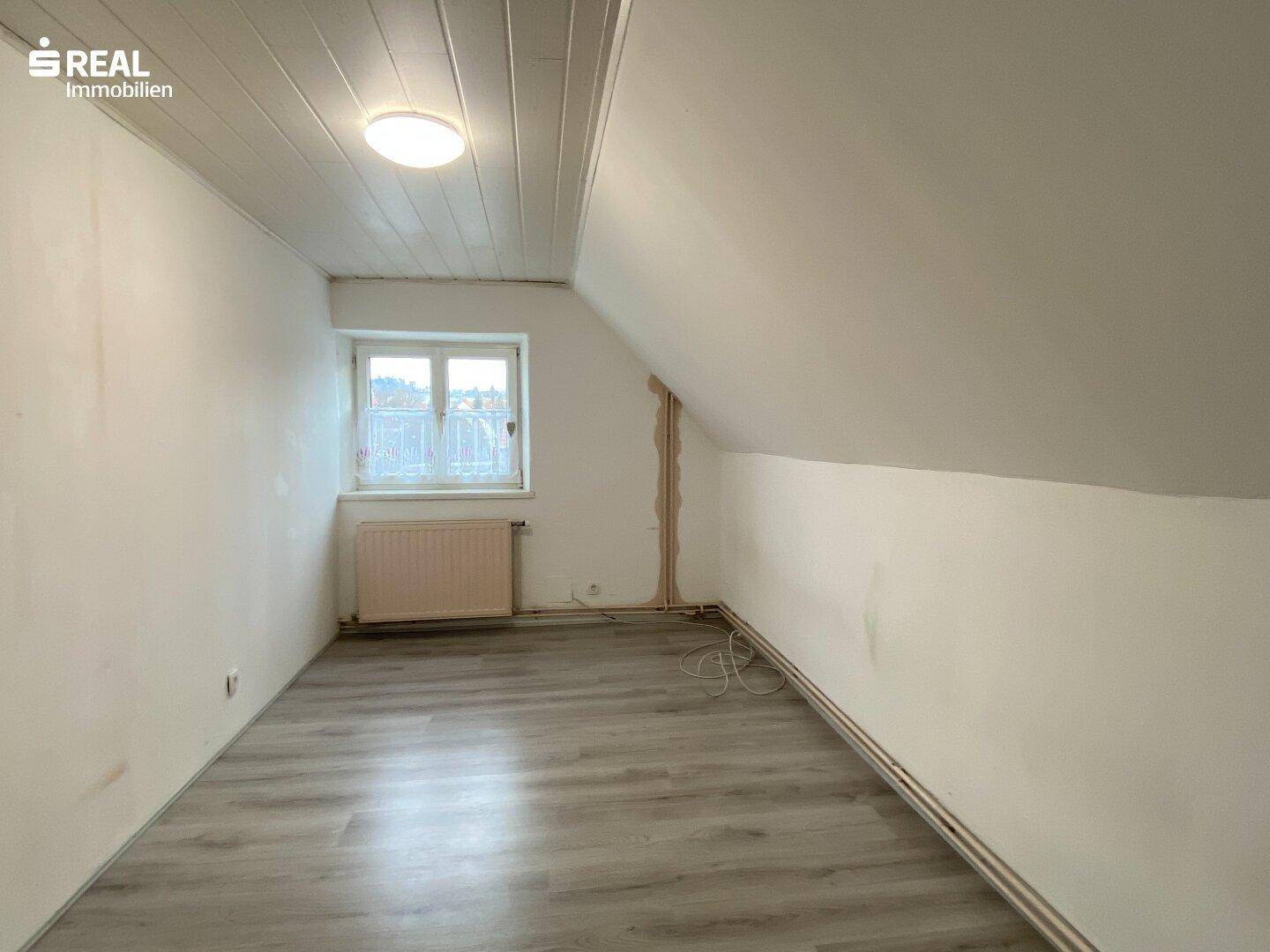 Helles Zimmer mit Dachschräge, Laminatboden und Blick nach draußen.