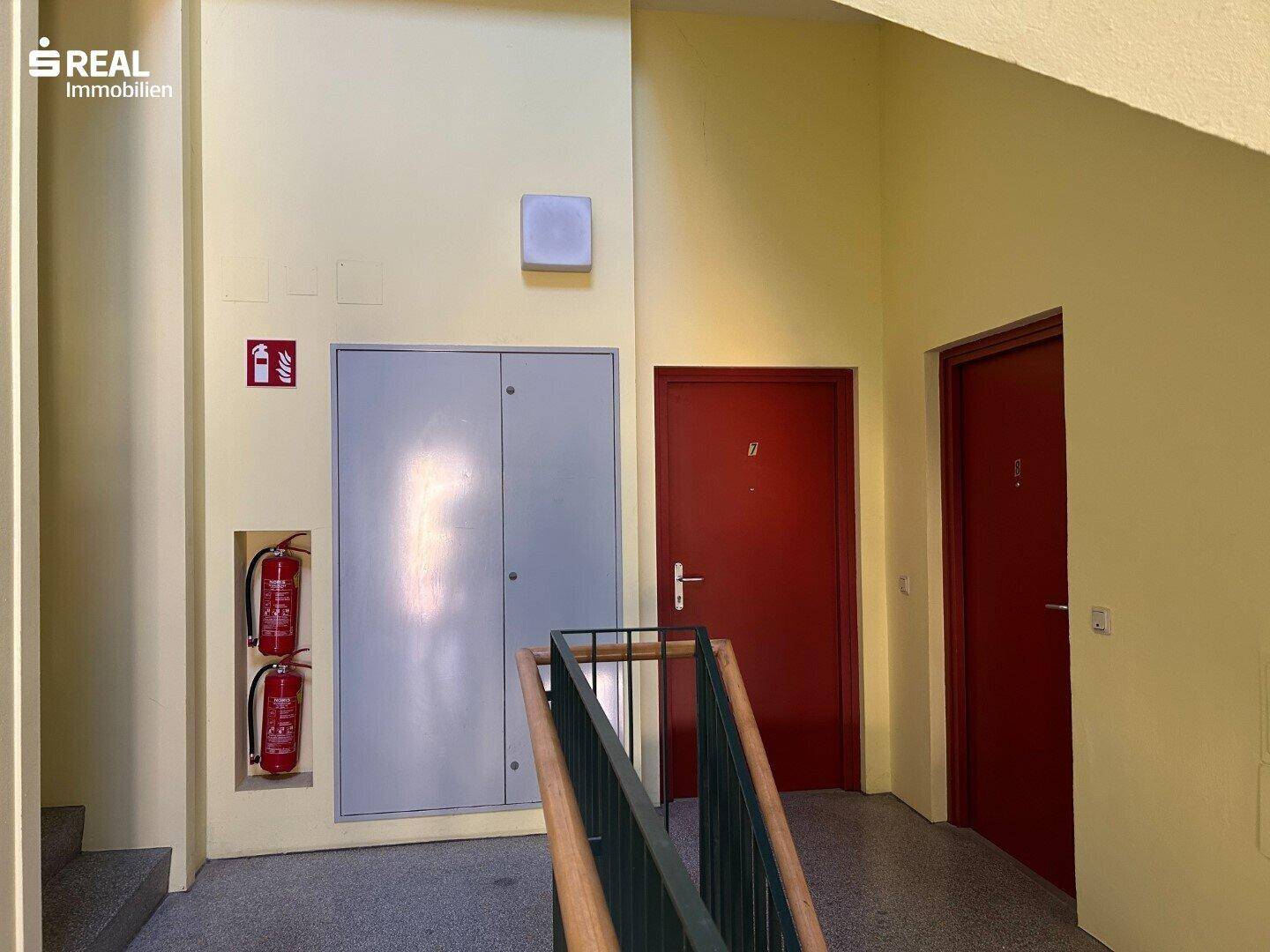 Treppenhaus mit gelben Wänden und roten Türen, ausgestattet mit Feuerlöschern.