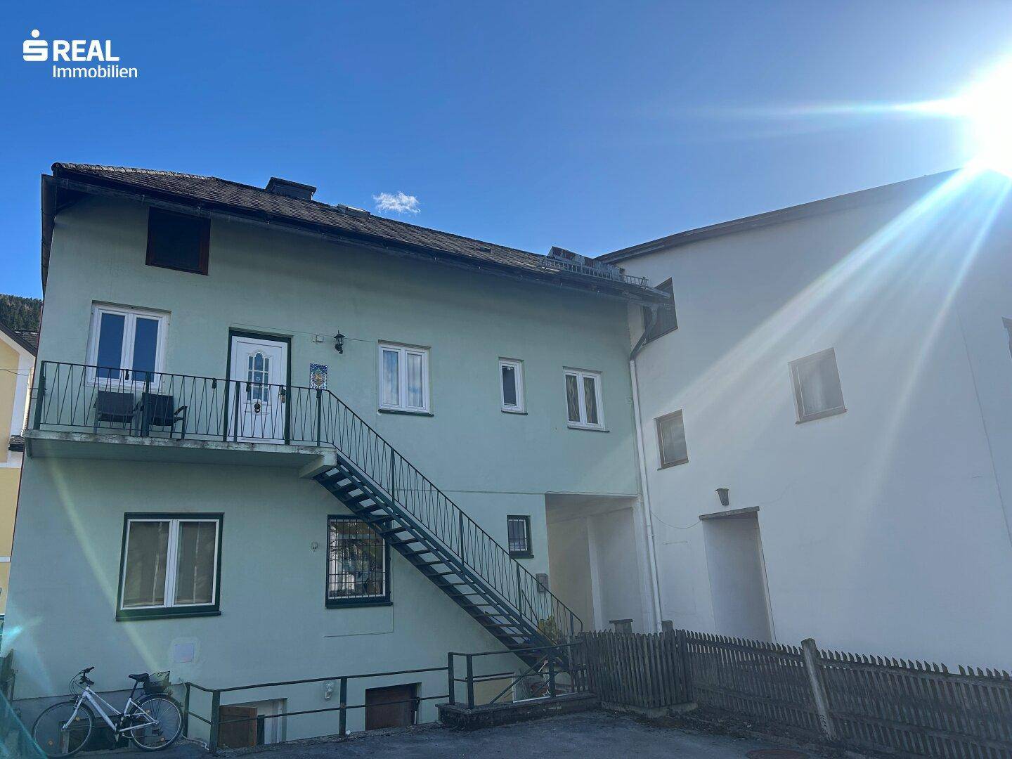 Rückseite eines Gebäudes mit externer Metalltreppe und einem kleinen Balkon unter strahlendem Sonnenschein.