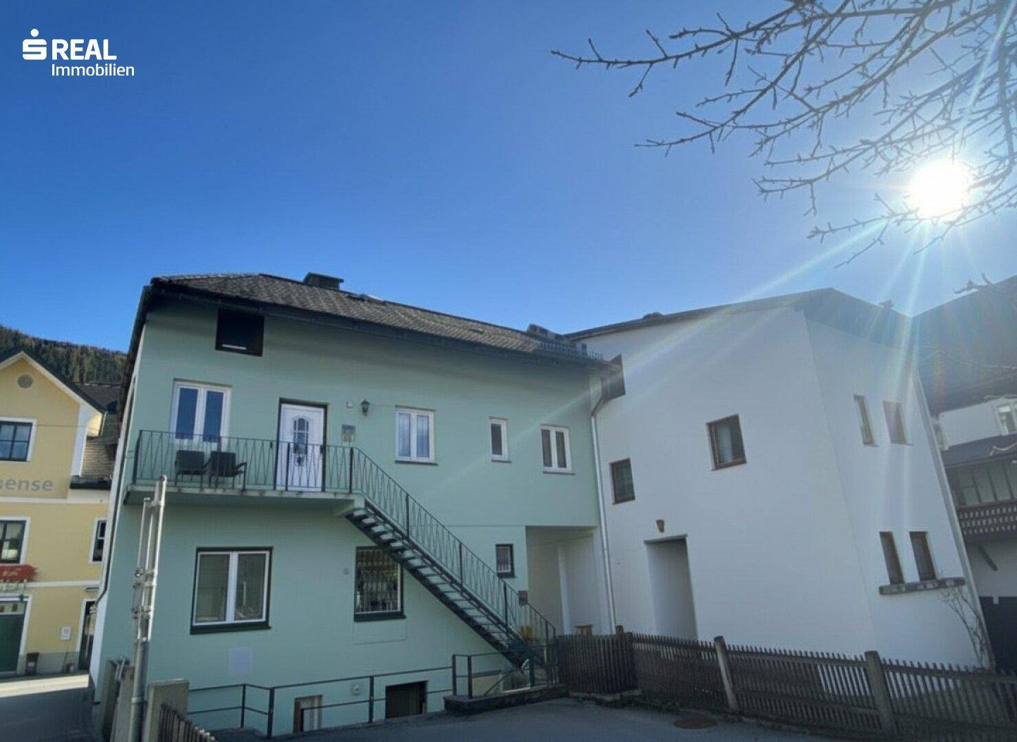 Gebäuderückseite mit Außentreppe und kleinem Balkon, umgeben von weiteren Häusern und viel Sonne.