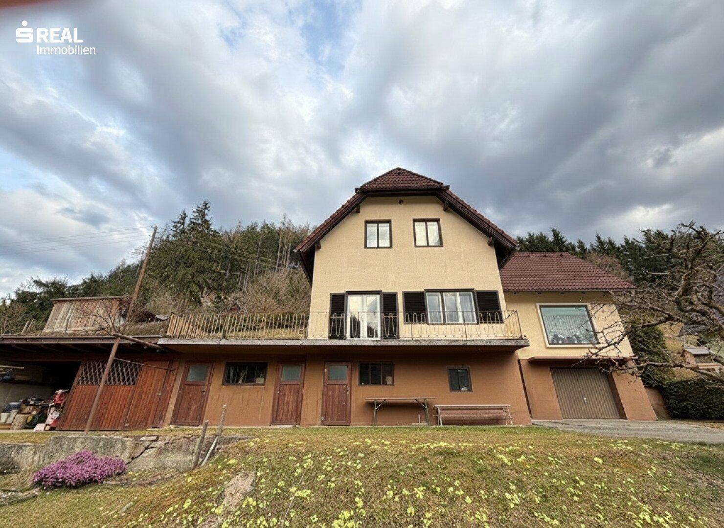Vorderansicht des Hauses mit Balkon und Garage, umgeben von grüner Natur und Hügeln.