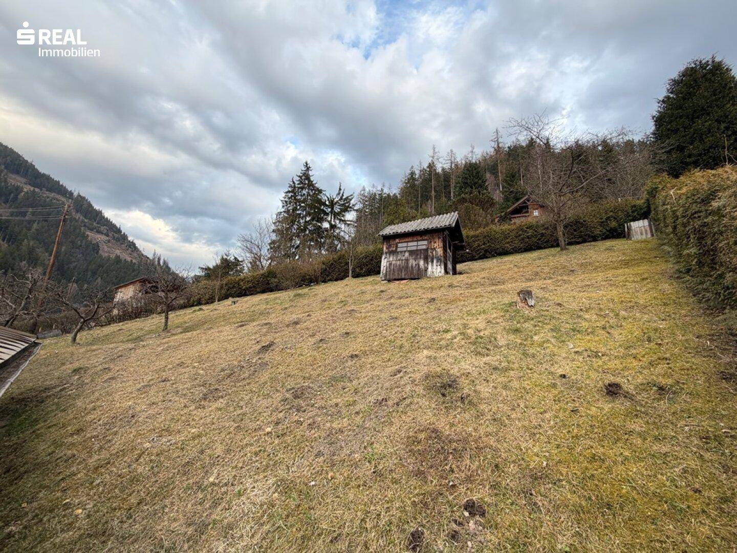Weitläufiger Gartenbereich mit einem kleinen Holzschuppen und Blick auf die bewaldeten Hügel.