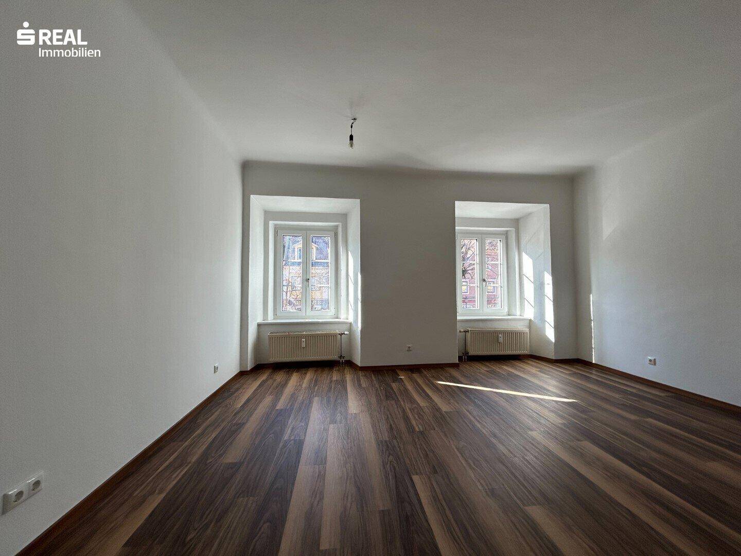 Geräumiger, heller Raum mit zwei Fenstern und dunklem Laminatboden.