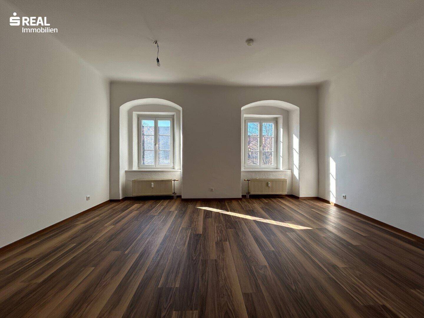 Geräumiger, heller Raum mit zwei Fenstern und dunklem Laminatboden.