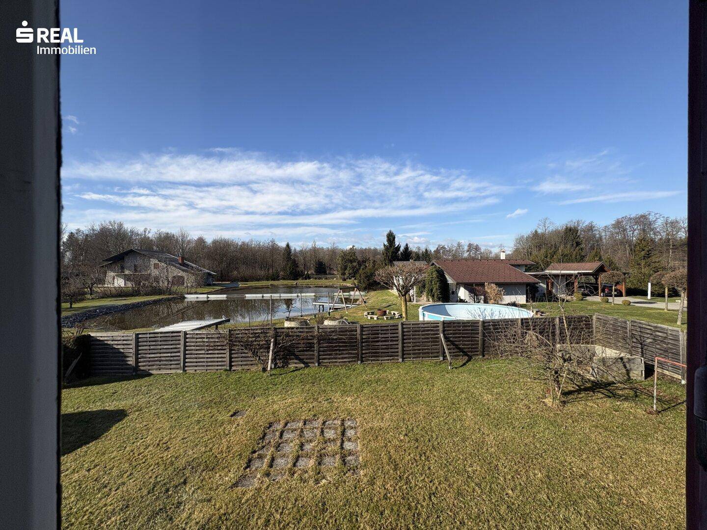 Weitläufiger Blick über den Garten mit Teich und Pool in der Ferne unter blauem Himmel.