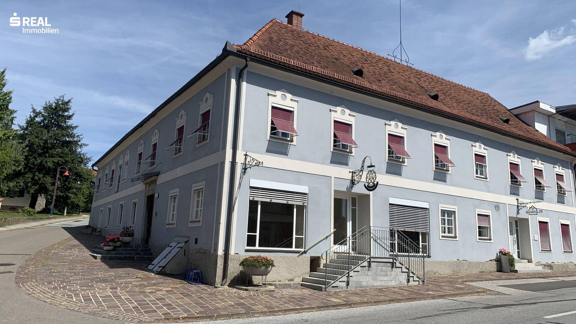 Eckansicht eines historischen Gebäudes mit hellblauer Fassade, roten Dachziegeln und vielen Fenstern.