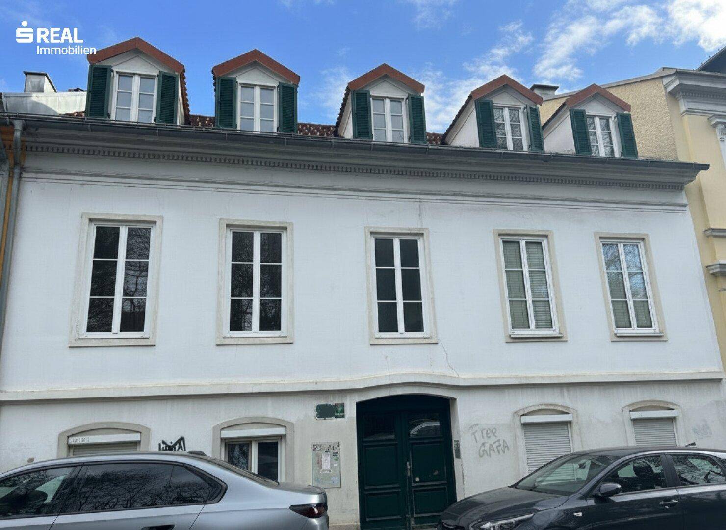 Nahaufnahme der historischen Fassade mit weißen Fenstern, grünen Fensterläden und Mansarddach.