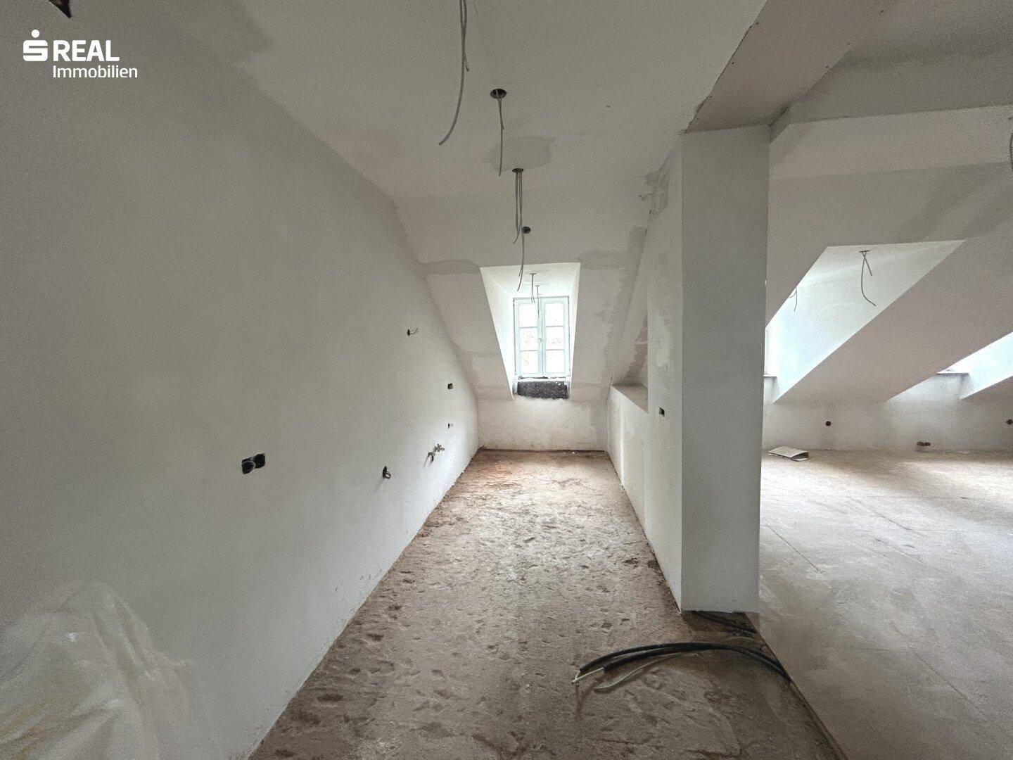 Langer Dachgeschossraum in Renovierung mit schrägen Wänden und einem Dachfenster.