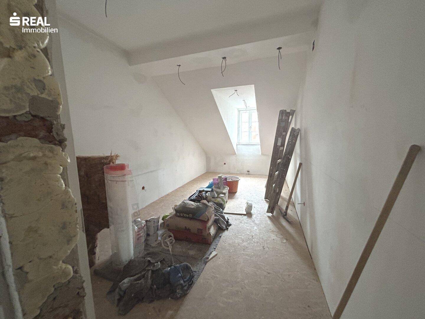 Dachgeschossraum in Renovierung mit Baumaterialien, schrägen Wänden und einem Dachfenster.