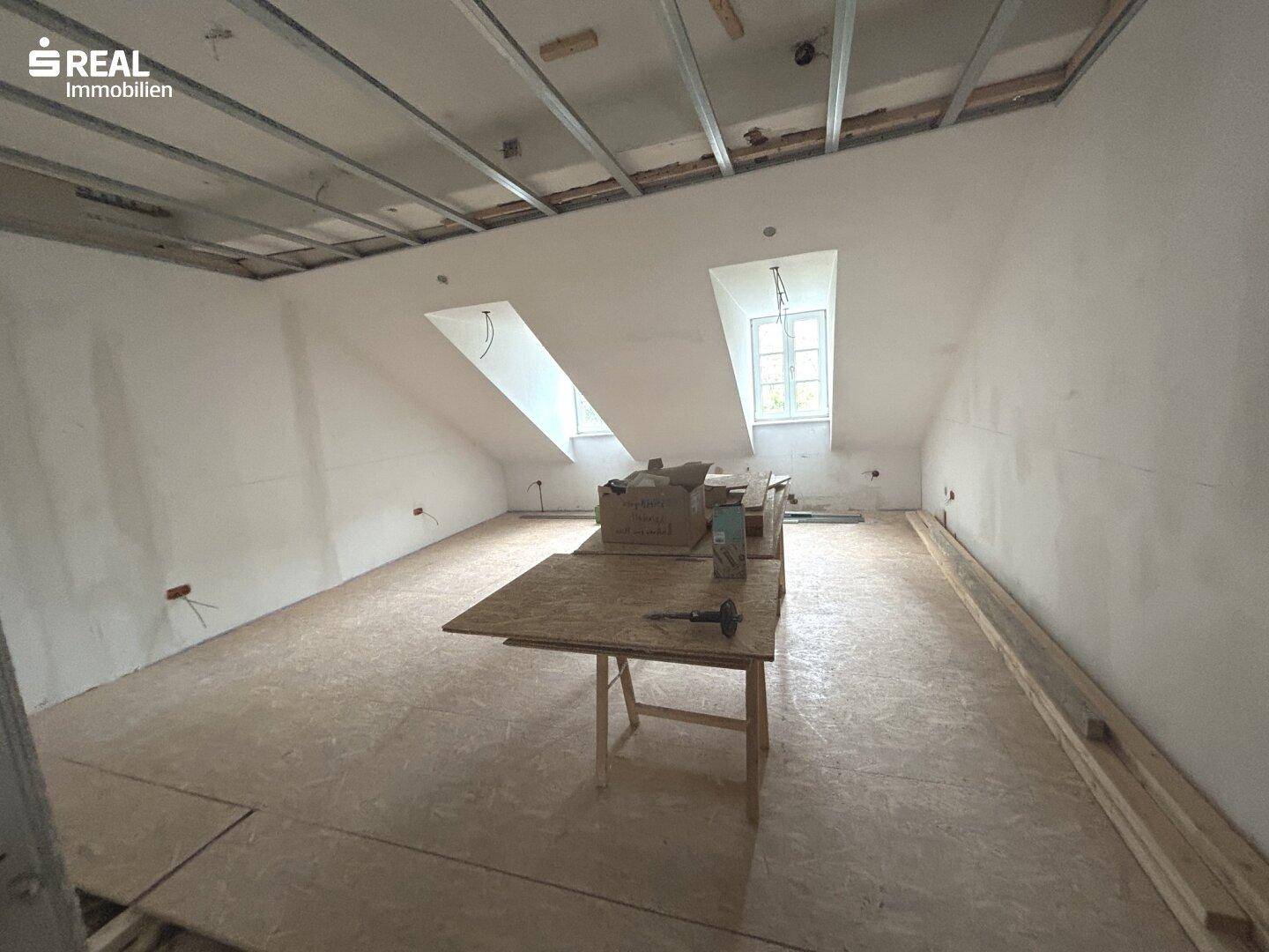 Dachgeschossraum in Renovierung mit sichtbarer Deckenkonstruktion, schrägen Wänden und Dachfenstern.