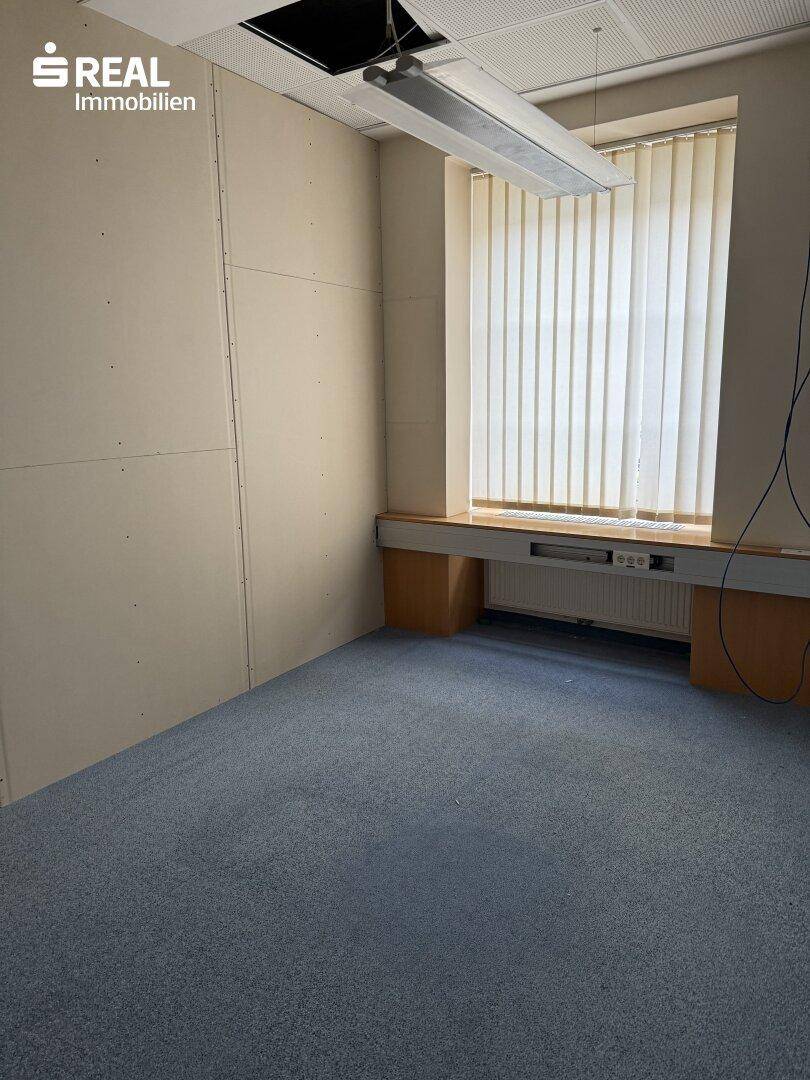 Büro mit Teppichboden, Fenster mit Jalousien und einer langen Arbeitsfläche.