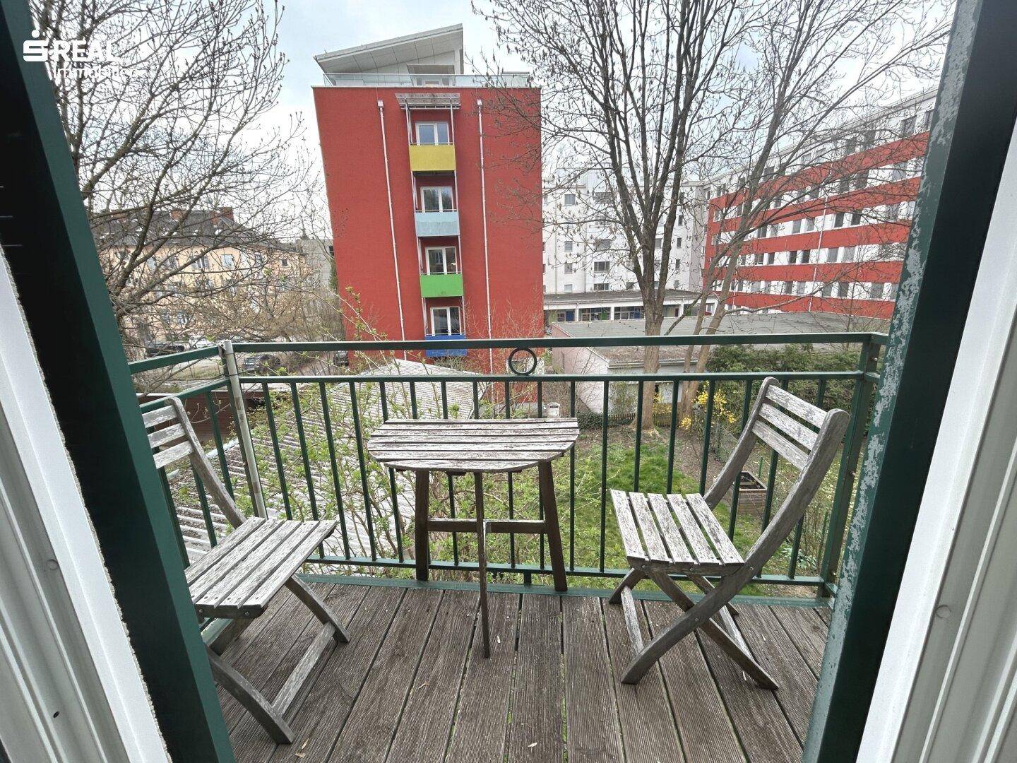 Kleiner Balkon mit Holzboden und Gartenmöbeln, umgeben von Bäumen und Gebäuden.