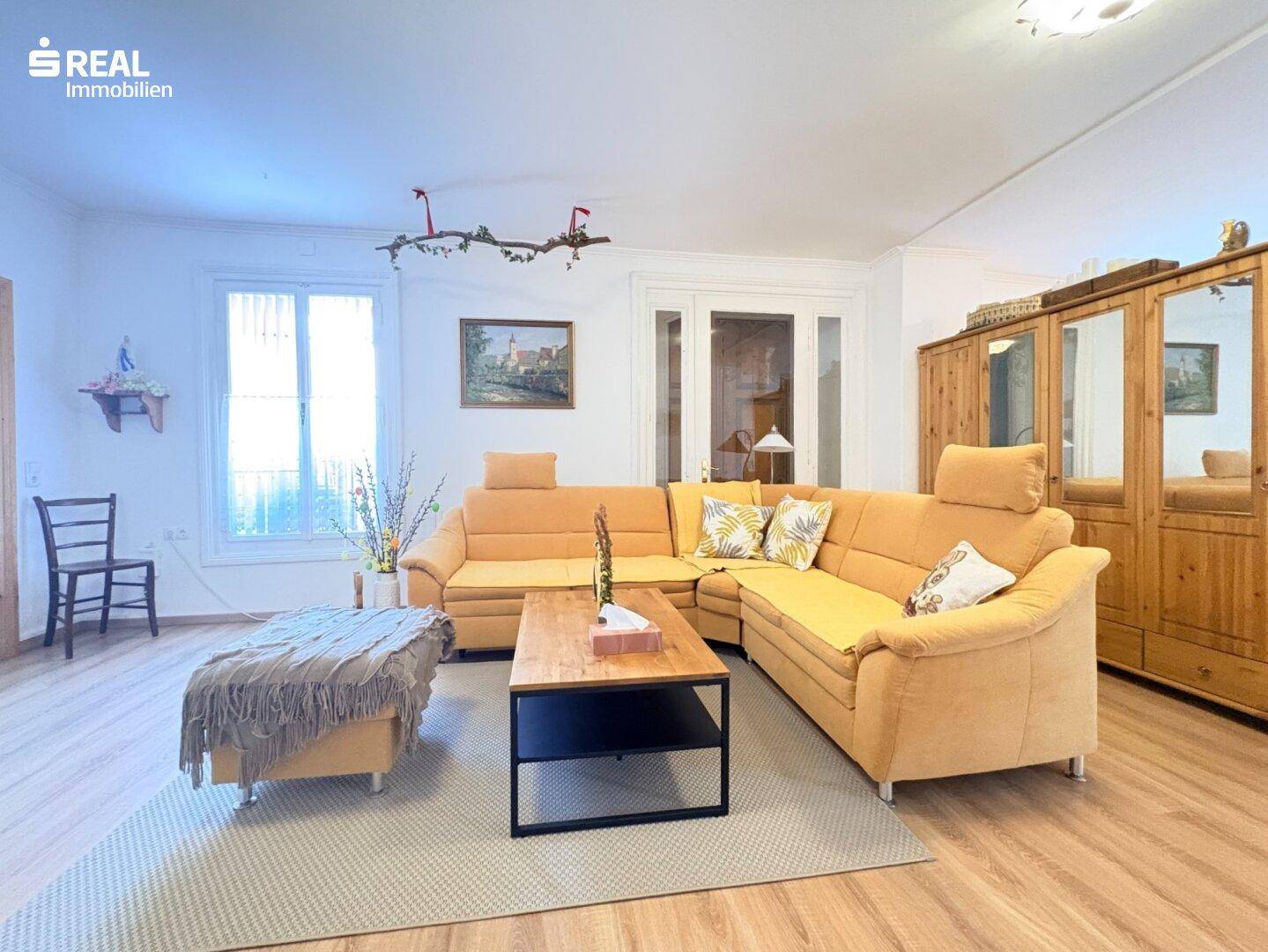 Helles Wohnzimmer mit gelbem Ecksofa, Holzboden und einem großen Holzschrank.