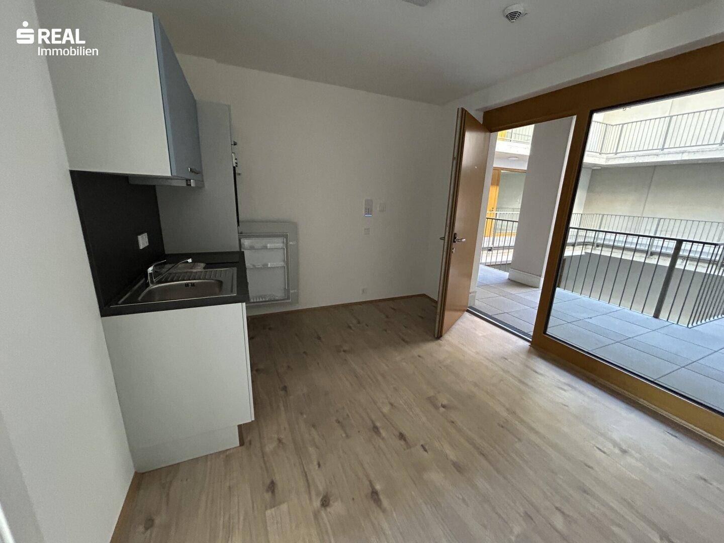 Wohnbereich mit integrierter Küchenzeile, Laminatboden und Zugang zu einem Balkon.