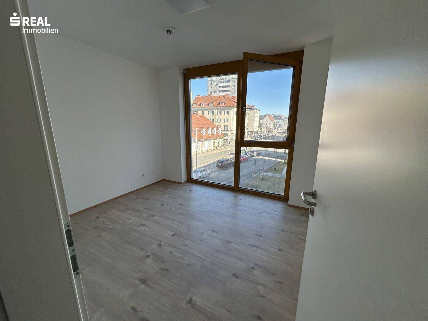 Helles, leeres Zimmer mit Laminatboden und großem Fenster mit Stadtblick.