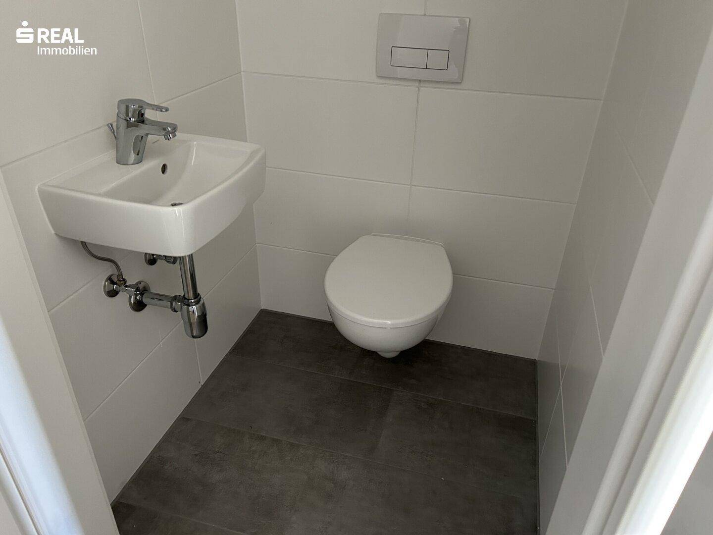 Separates Gäste-WC mit wandhängender Toilette, kleinem Waschbecken und dunklen Bodenfliesen.