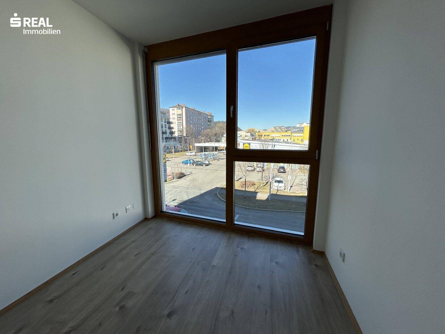 Helles Zimmer mit Laminatboden und breitem Fenster mit Blick auf den Parkplatz.