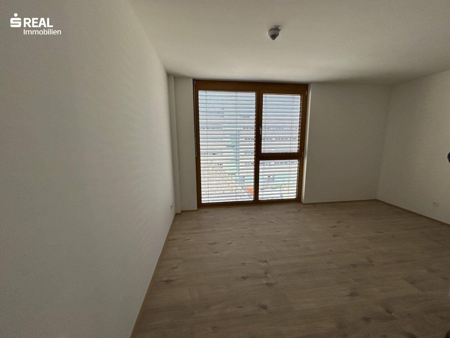 Helles Zimmer mit Laminatboden und Fenster mit integrierten Jalousien.