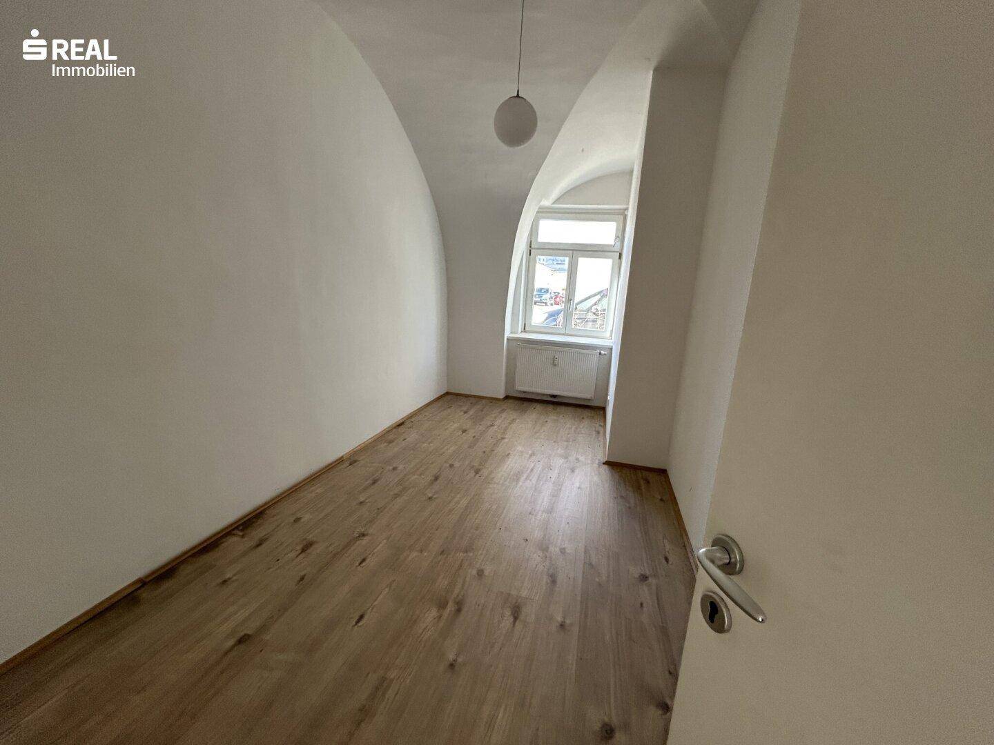 Helles Zimmer mit Laminatboden, Gewölbedecke und Blick aus dem Fenster.
