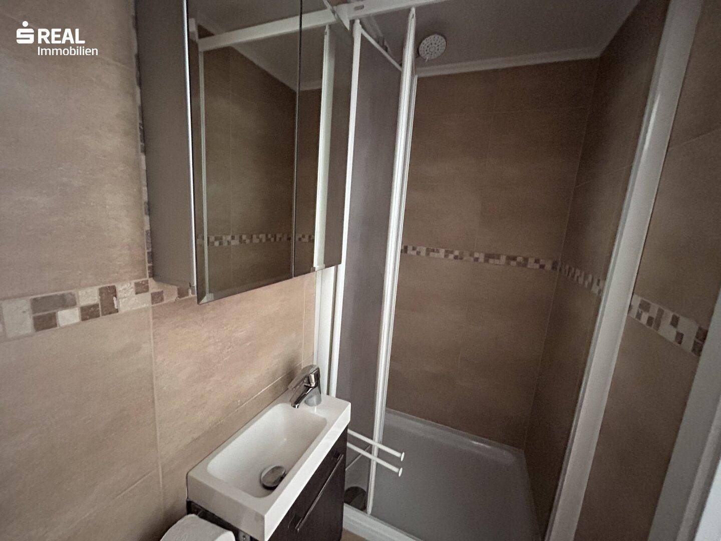 Kleines Badezimmer mit Duschkabine, Waschbecken und Spiegelschrank, komplett gefliest.