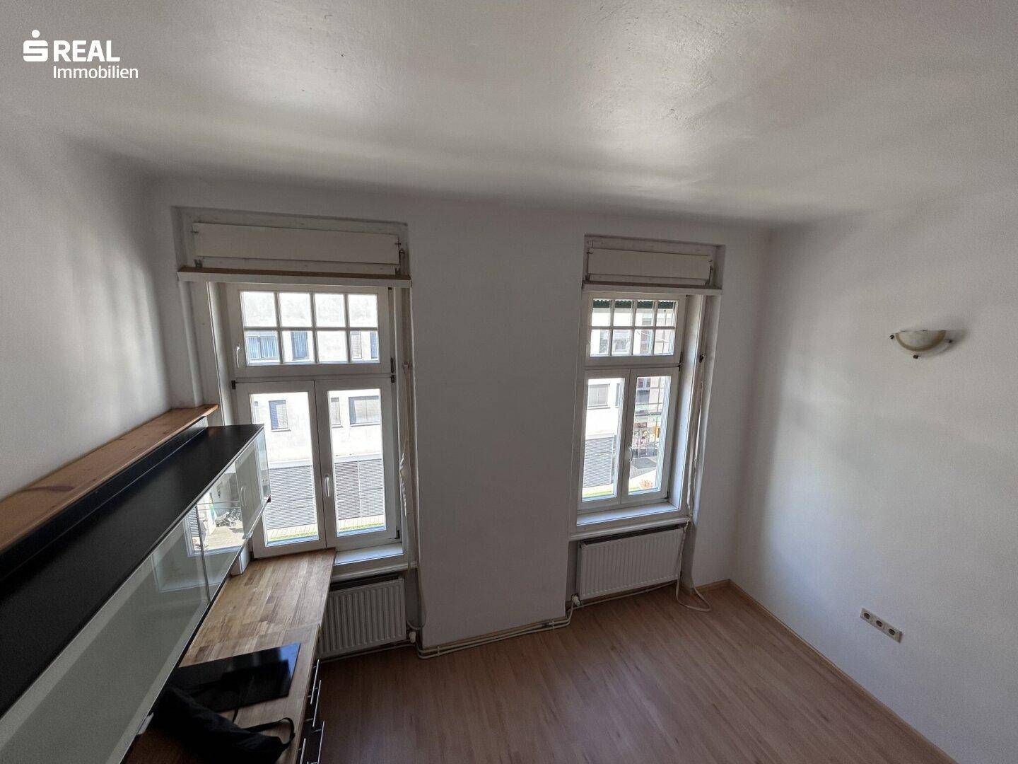 Geräumiger Wohnbereich mit zwei großen Fenstern, die viel Tageslicht hereinlassen.