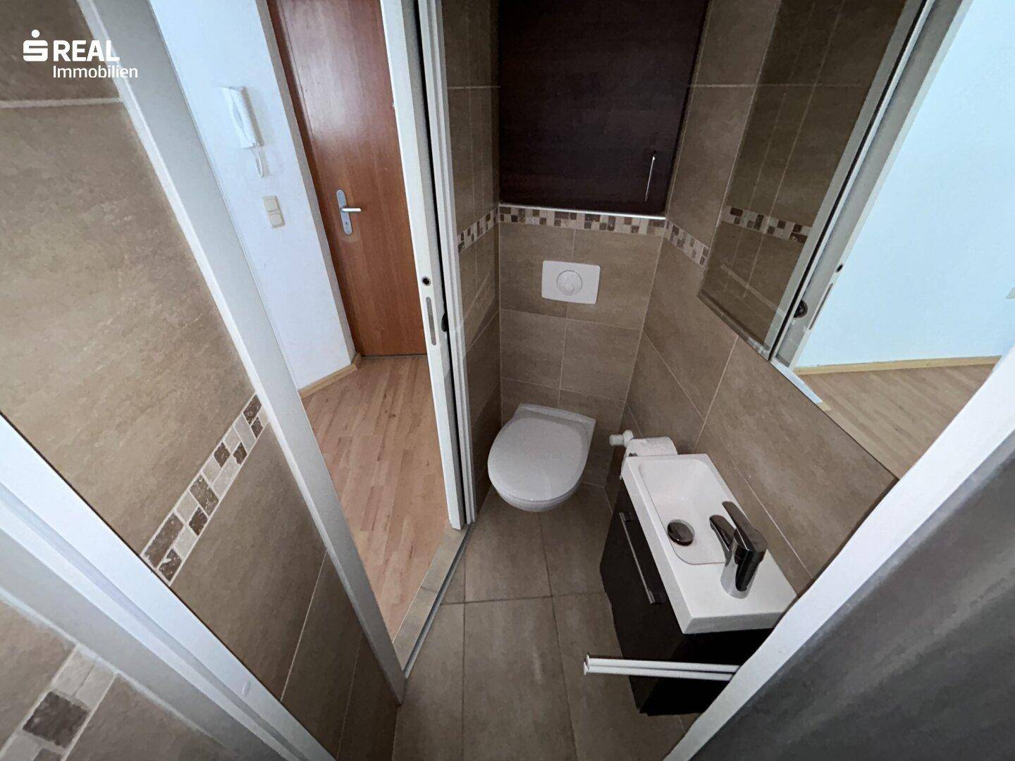 Kompaktes WC mit Wand-WC, kleinem Waschbecken und beigefarbenen Fliesen.