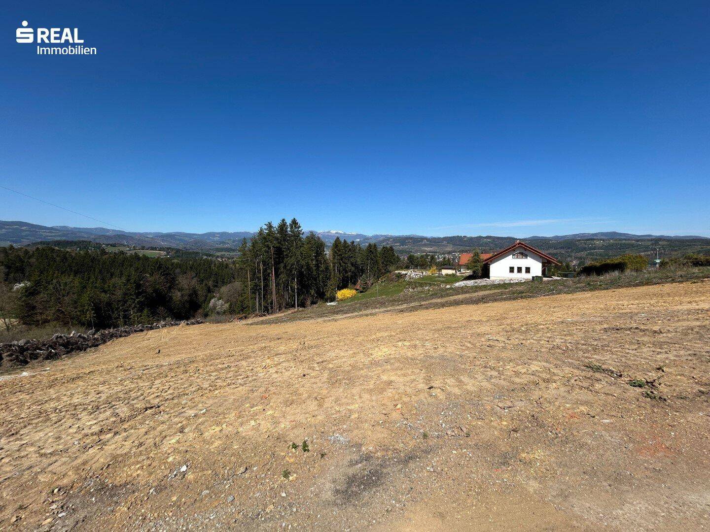 Sonniges Grundstück mit Aussicht auf ein Wohnhaus, Wald und die Bergkette am Horizont.