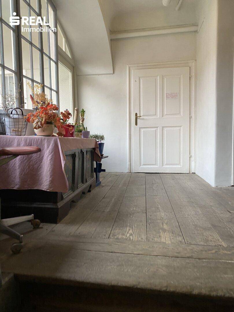 Raum mit breiten Holzdielen, großen Fenstern und einfacher Einrichtung in einem historischen Gebäude.