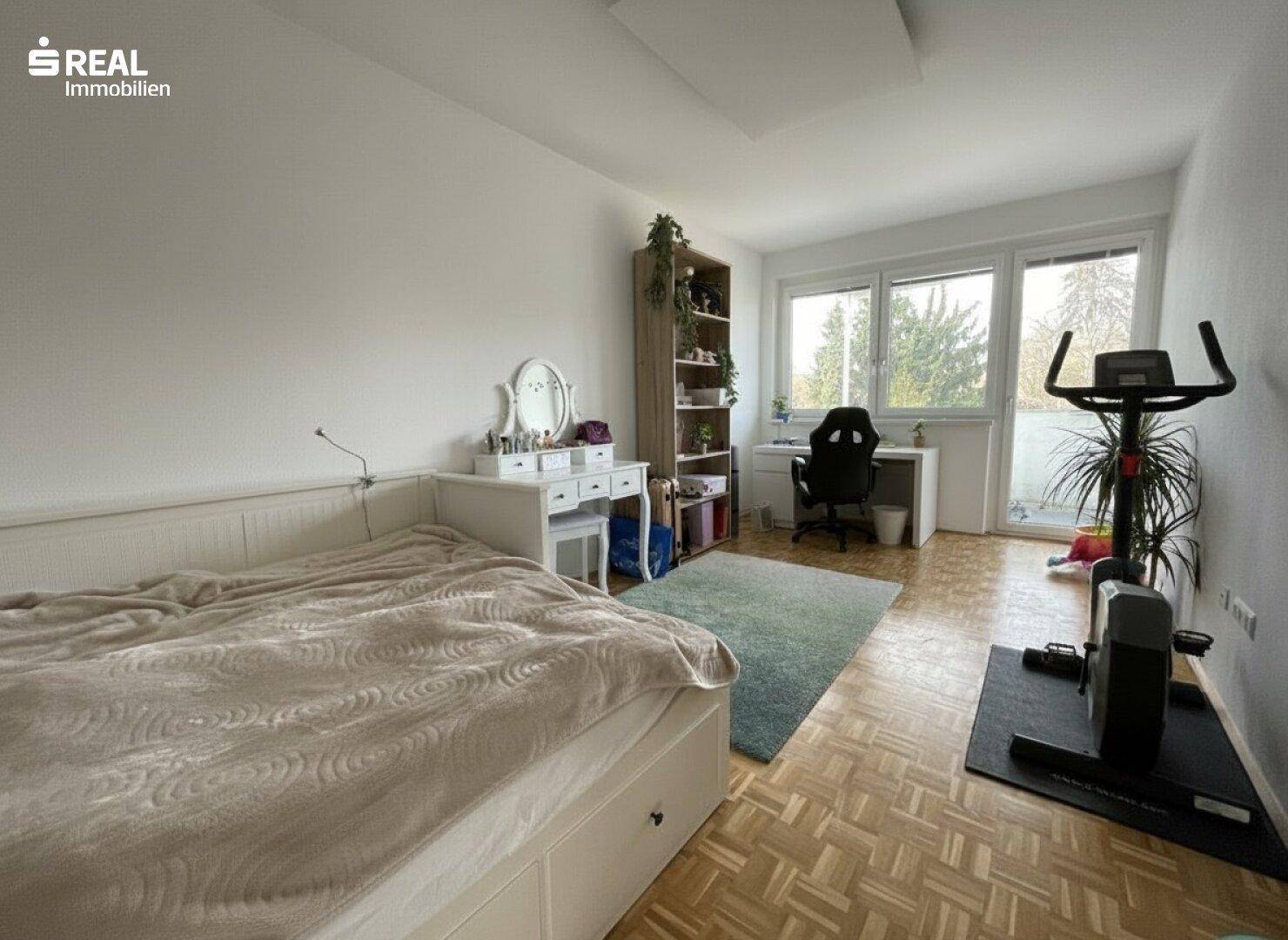 Helles Schlafzimmer mit Schreibtisch, Sportgerät und Zugang zu einem Balkon.