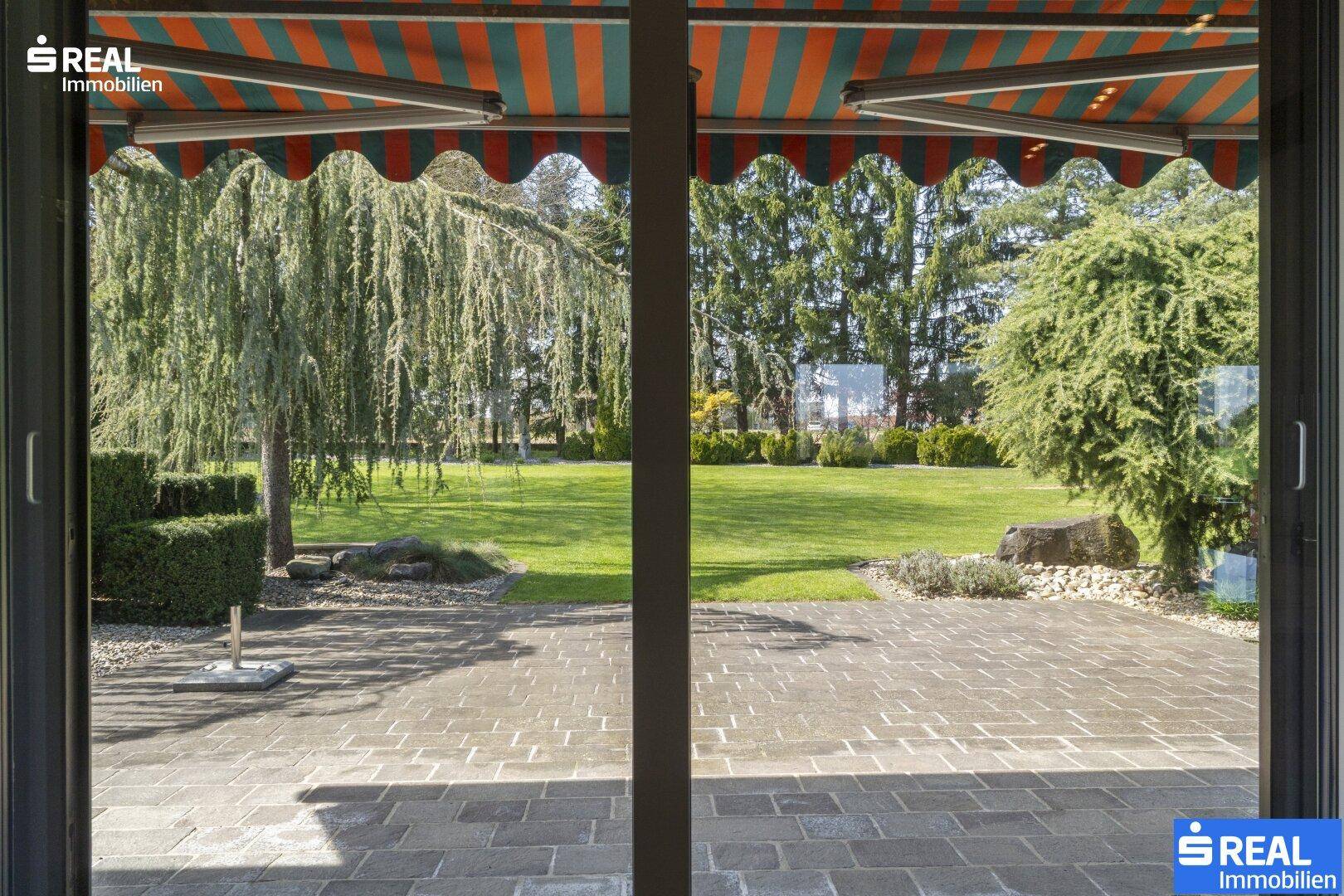 Blick von innen auf eine gepflasterte Terrasse mit Markise und den weitläufigen, grünen Garten.