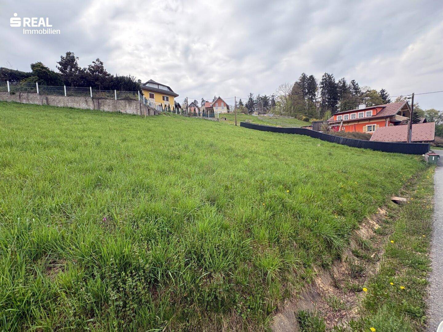 Weitläufiges grünes Grundstück mit leichter Neigung und Häusern in der Ferne unter bewölktem Himmel.