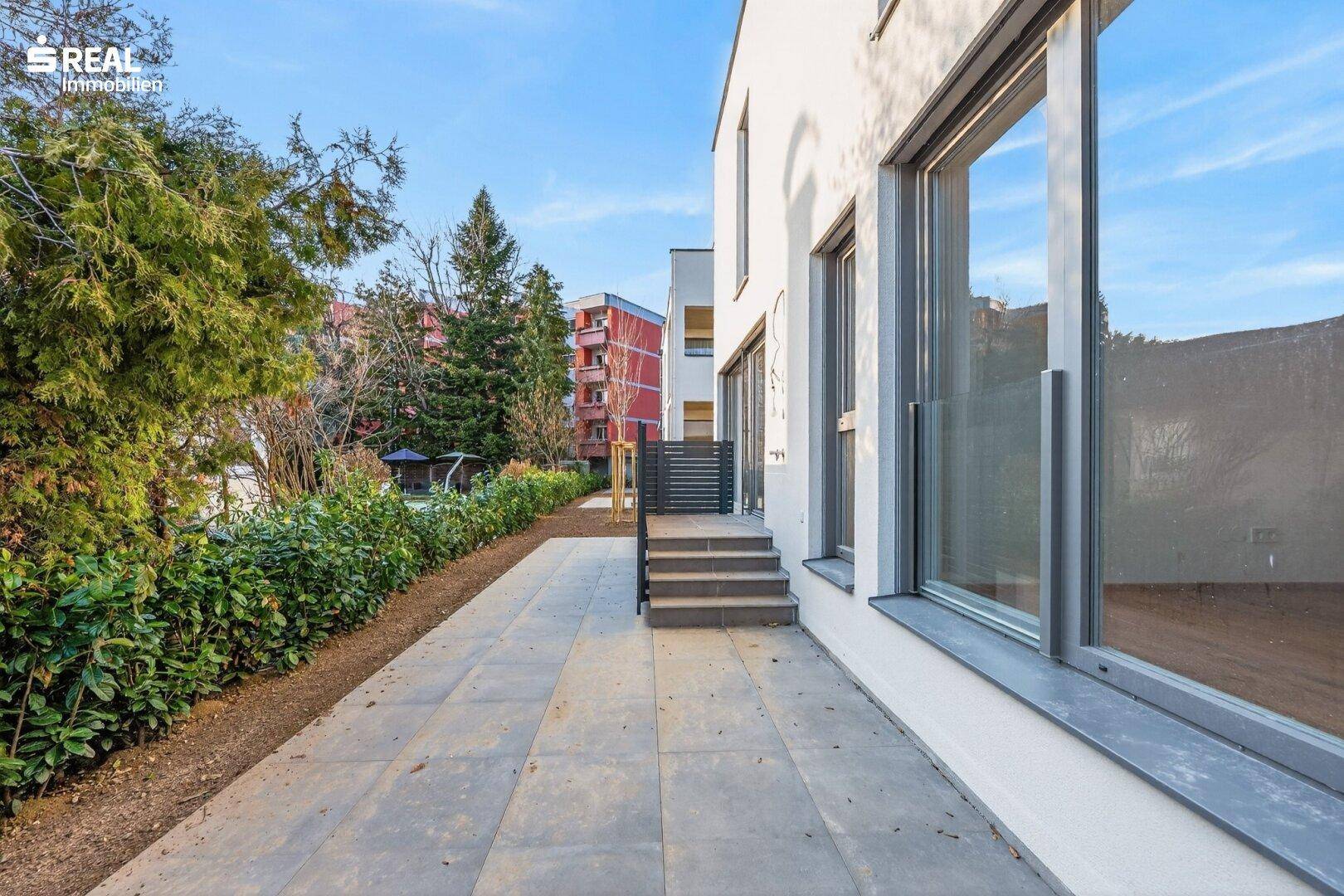 Langer Terrassenweg aus Steinplatten entlang der modernen Hausfassade mit viel Grün.