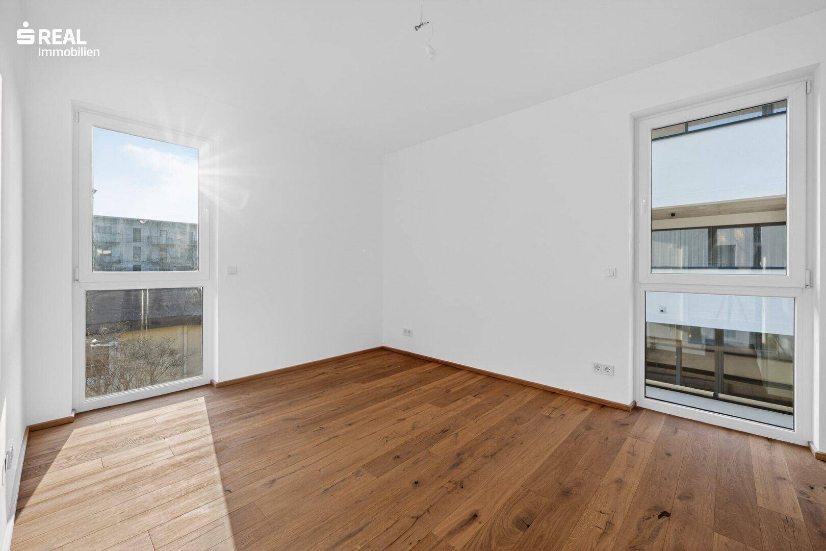 Sonniger Raum mit zwei großen Fenstern und Parkettboden, ideal als Schlaf- oder Arbeitszimmer.