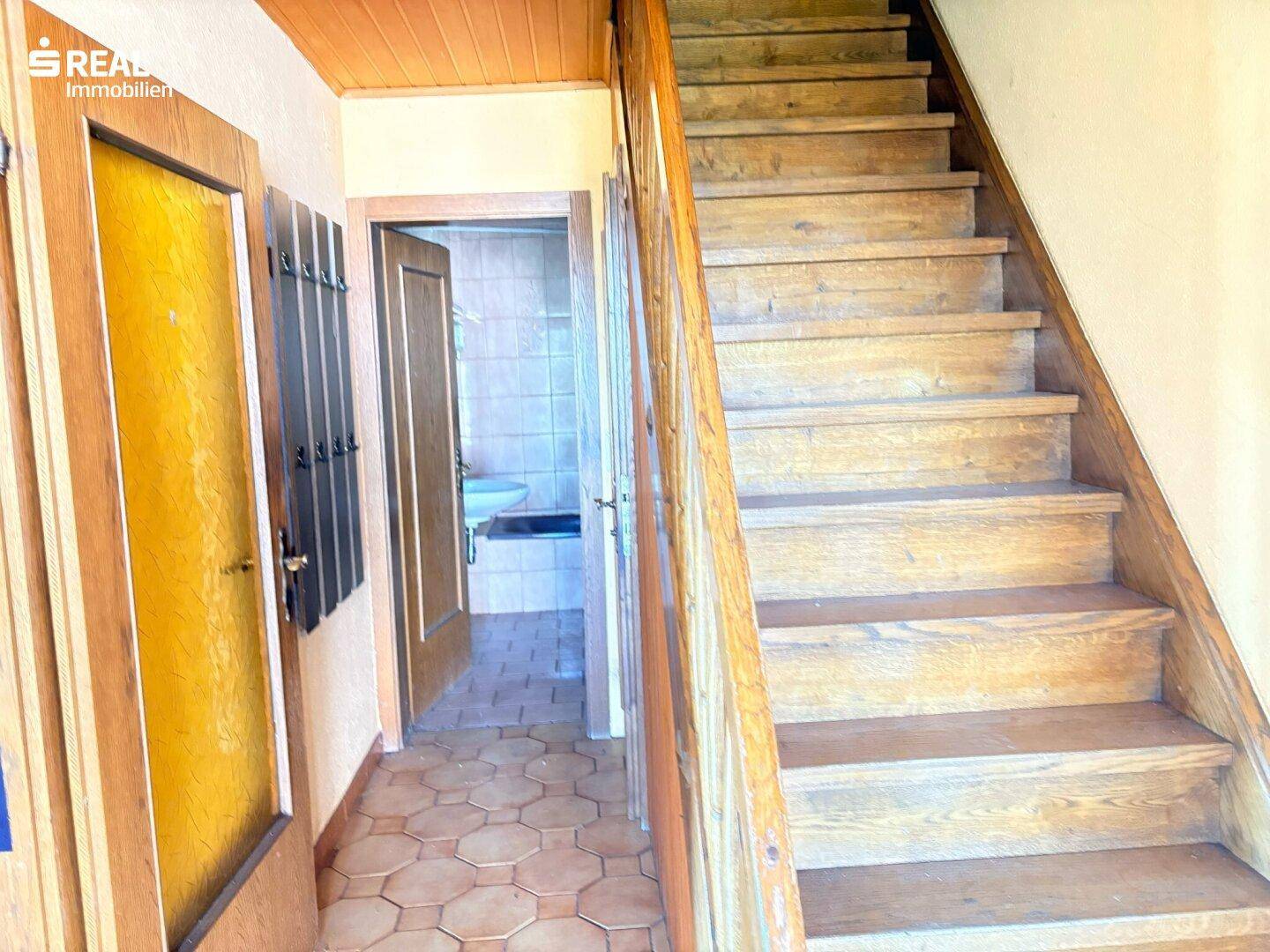 Hölzerne Treppe führt in die obere Etage, mit Fliesenboden und Blick auf ein Badezimmer.