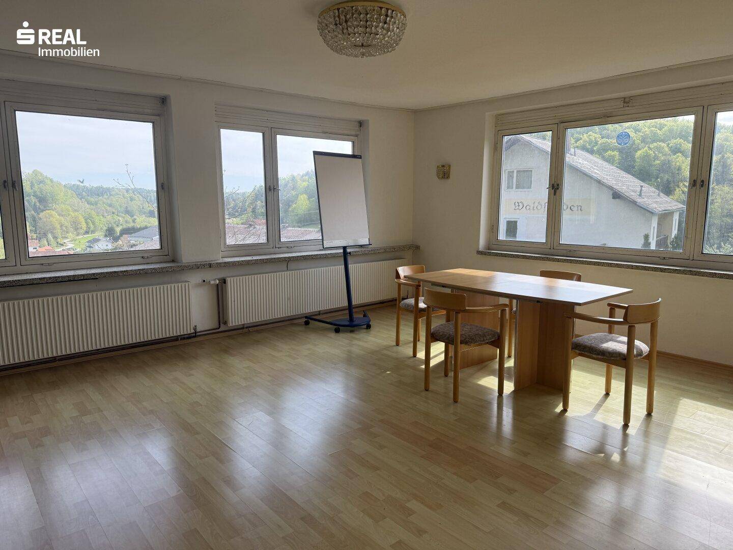 Heller Mehrzweckraum mit Holzboden, großen Fenstern und einem Konferenztisch, ideal für Besprechungen.