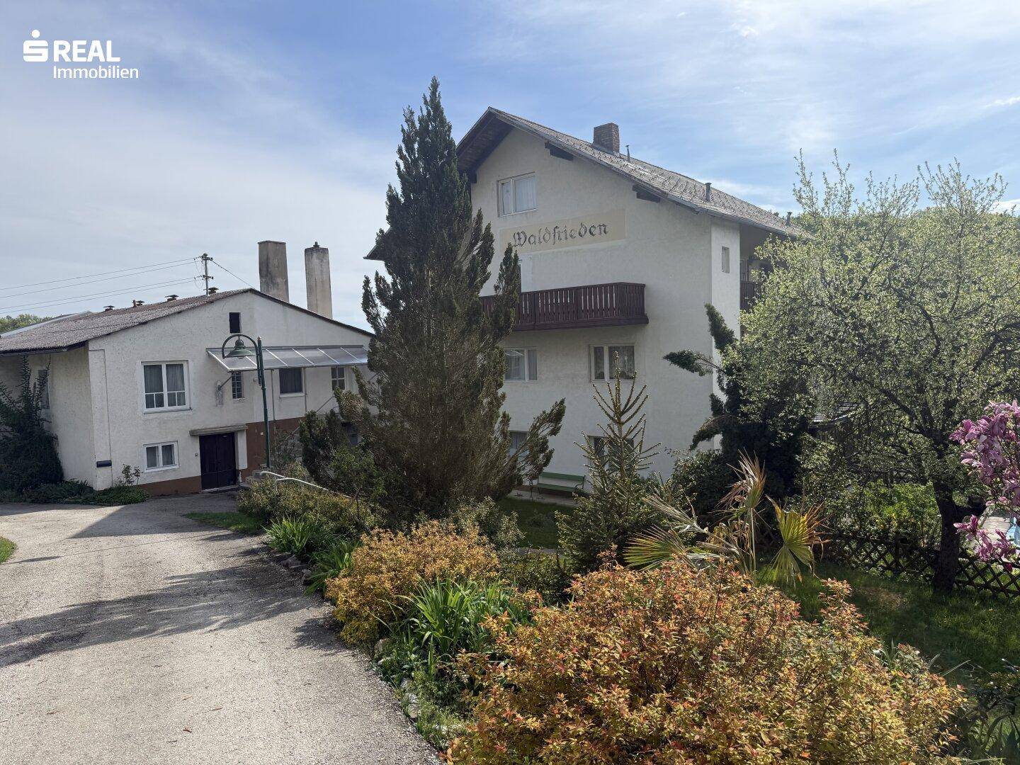 Weißes Gebäude mit traditioneller Architektur, Balkonen und einem gepflegten Vorgarten mit Bäumen.
