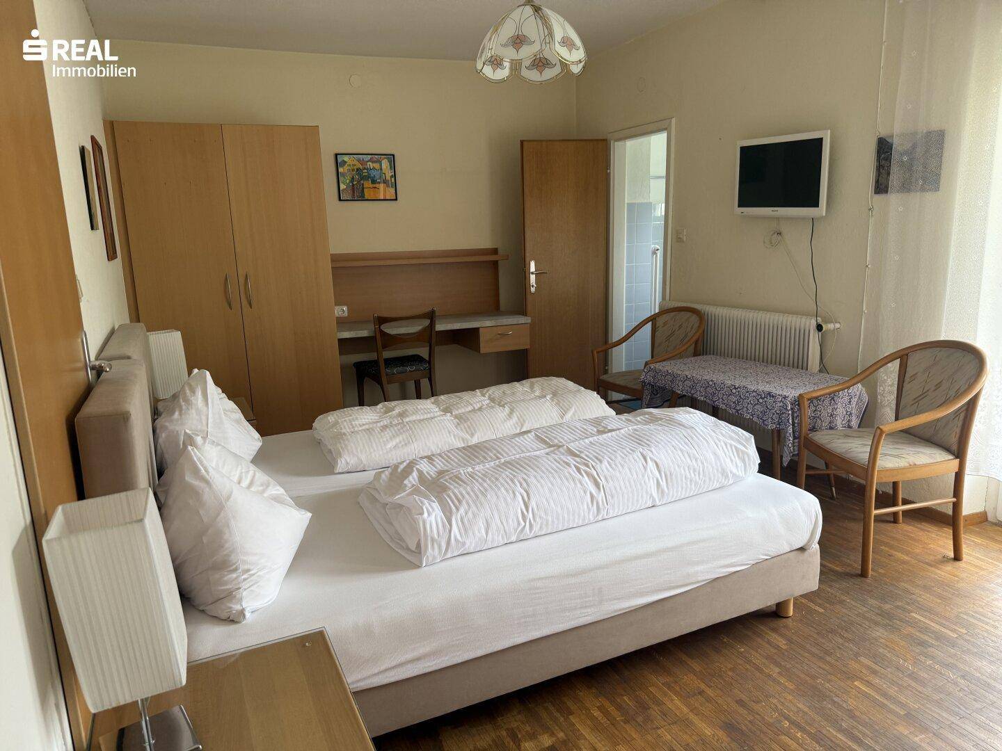 Einfaches Schlafzimmer mit zwei Betten, Schrank und einem kleinen Schreibtisch, mit Blick auf die Umgebung.