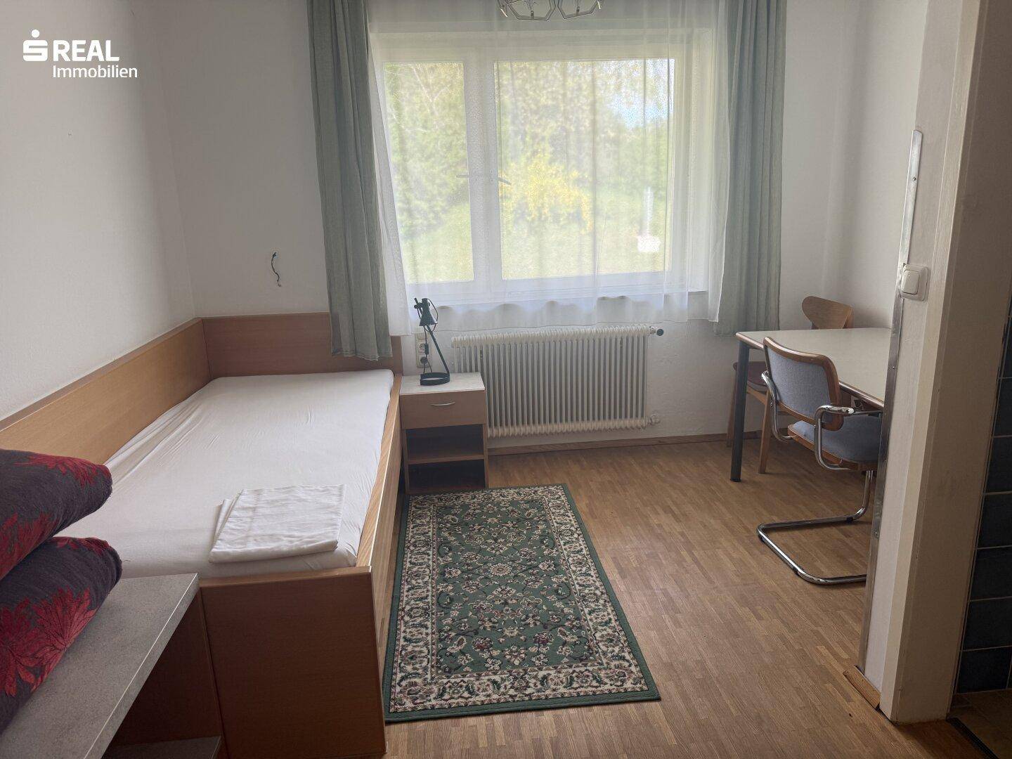 Helles Einzelzimmer mit Holzboden, einem Bett und einem Schreibtisch, gut beleuchtet durch ein großes Fenster.