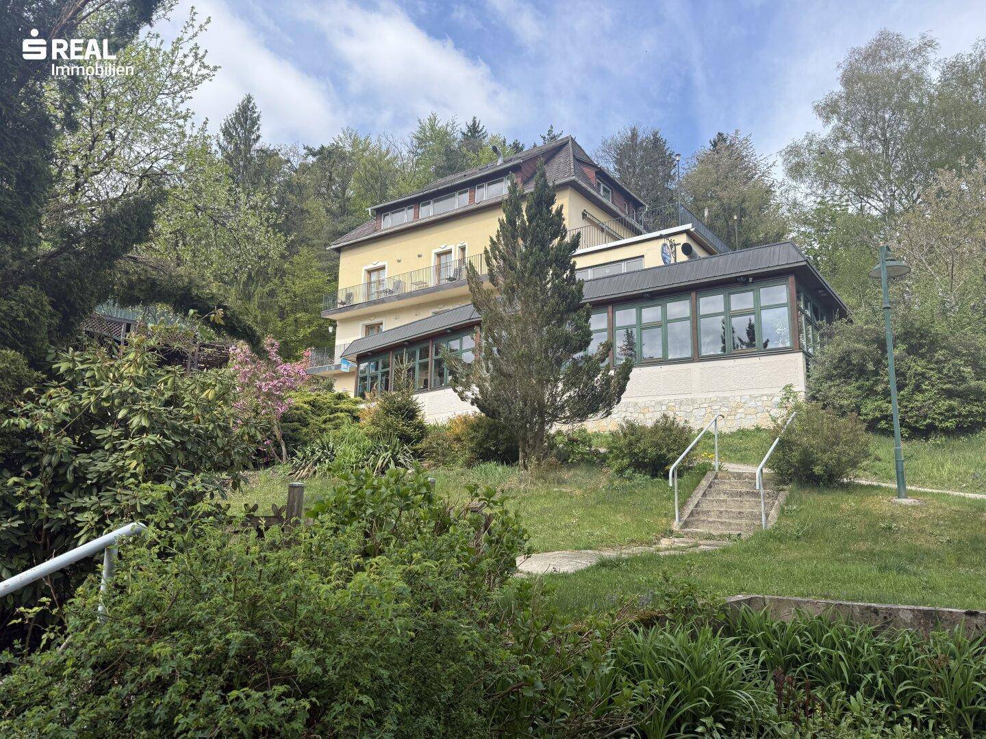 Mehrstöckiges Gebäude in Hanglage, umgeben von üppiger Vegetation und Bäumen unter blauem Himmel.