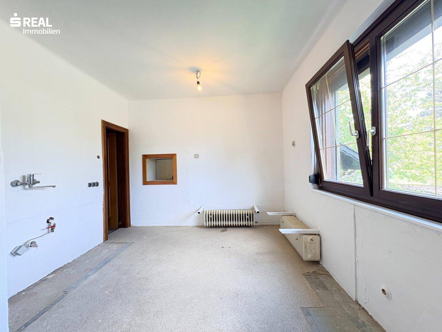 Leerer Küchenraum mit Fenster, Heizkörper und Anschlüssen für eine Einbauküche.