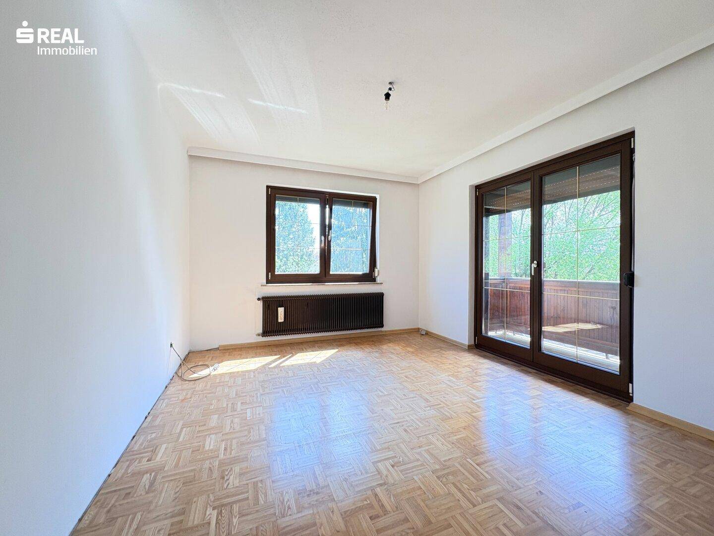 Helles Zimmer mit Parkettboden, großem Fenster und Zugang zu einem Balkon.