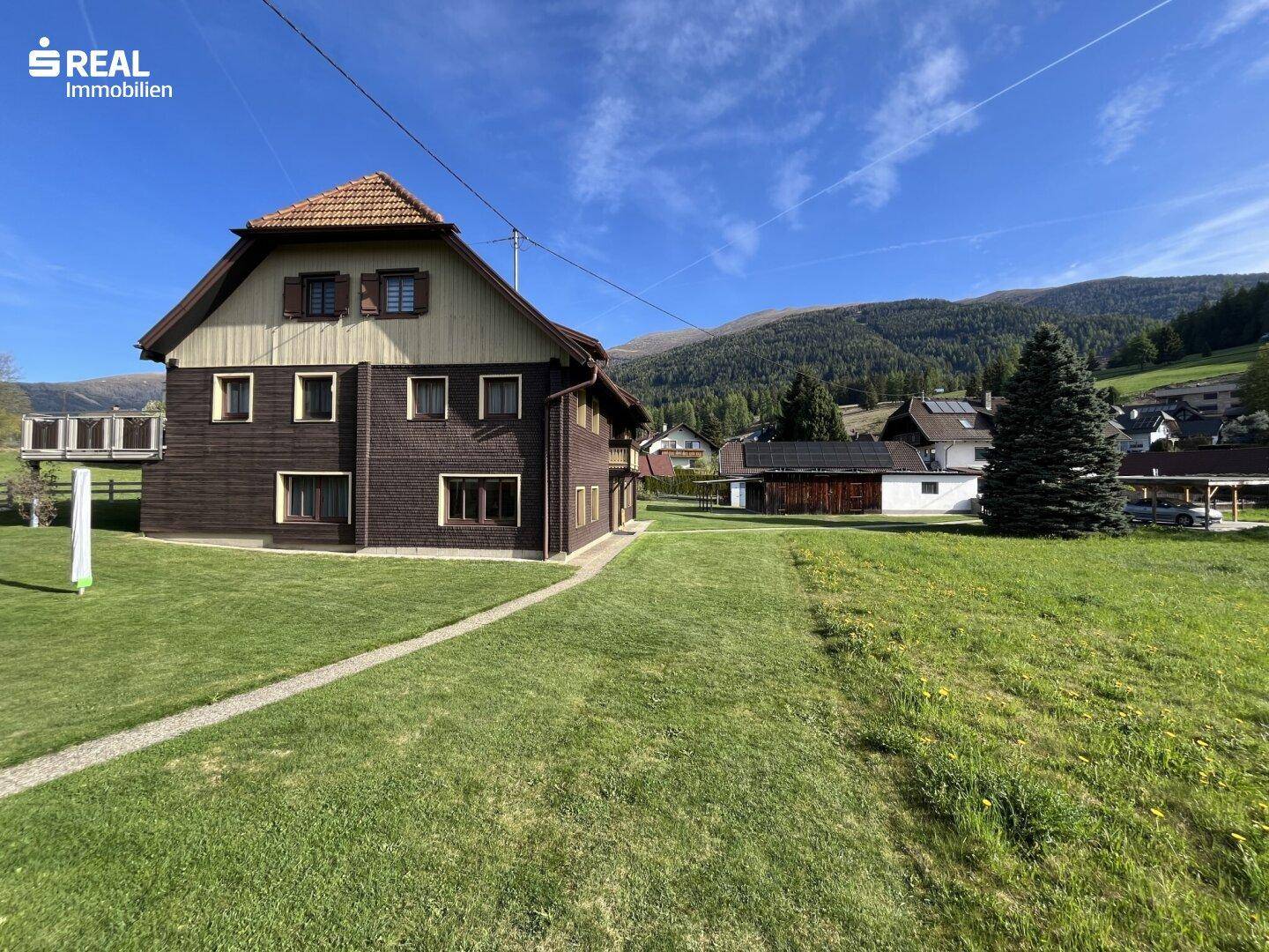 Traditionelles Wohnhaus mit Holzfassade und weitläufiger Rasenfläche vor einer Bergkulisse.
