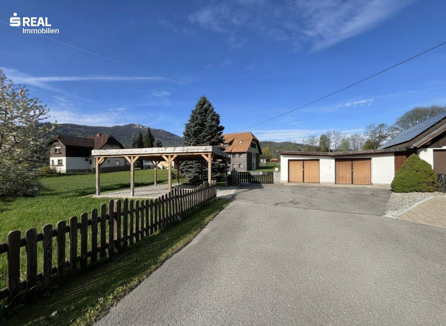 Zufahrt zum Anwesen mit Carport, Garage und Holzzaun, umgeben von grüner Landschaft.