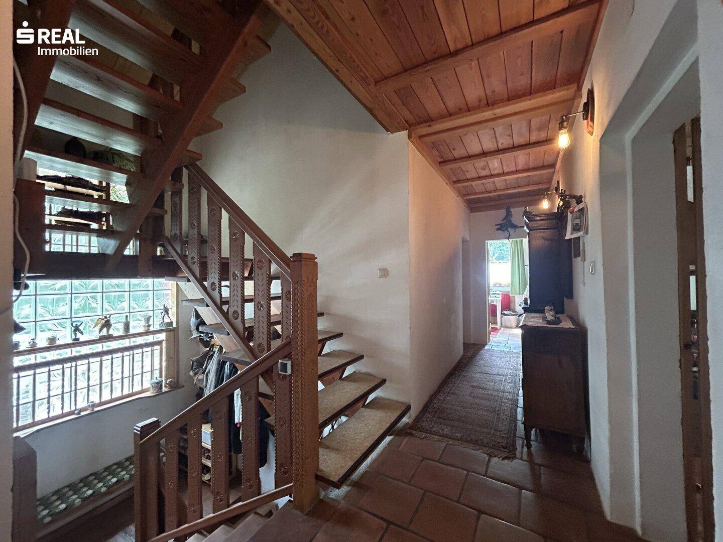 Rustikaler Flur mit Holztreppe, Holzbalkendecke und Terrakotta-Fliesenboden, der zu weiteren Räumen führt.